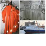 Oranžs ūdenslīdēju hidrotērps piekārts uz metāla pakaramajiem, fona attēlā redzams kuģis.